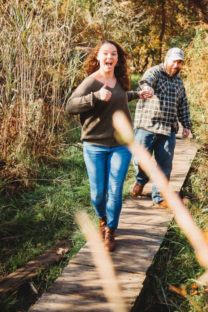 A man and woman who are handcuffed together run along a wooden footbridge over shallow water amongst reeds during their fall Haley Farm engagement session.