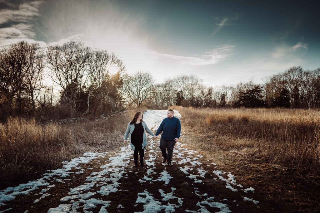 A woman and man walk and hold hands while looking at each other with a blue sky, golden grass, and snow remnants during their winter Haley Farm engagement session.