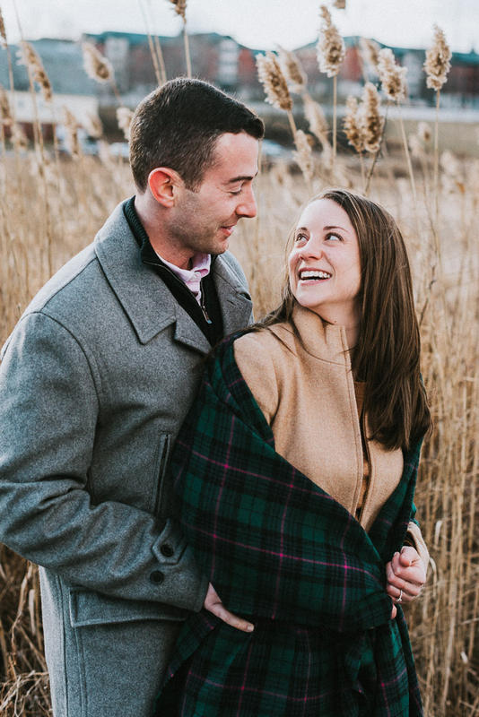 A man in a gray coat and woman in a tartan scarf stand in front of golden reeds during their UConn engagement session.