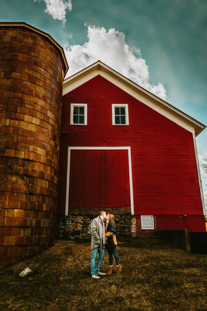 A couple embraces and kisses in front of Jacobson Barn during their UConn engagement session.