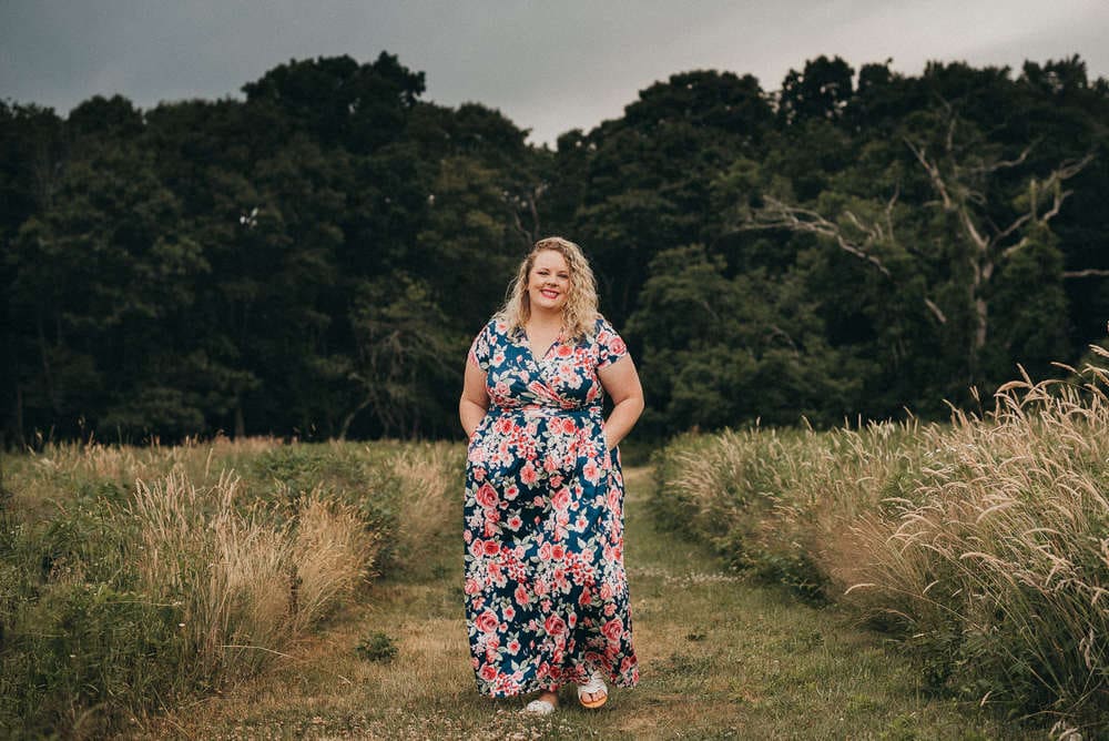 A woman in a blue and pink floral dress walks down a mowed path through a high grass field with deep green trees behind her during a faux southeastern Connecticut engagement session for a Connecticut wedding photography video.