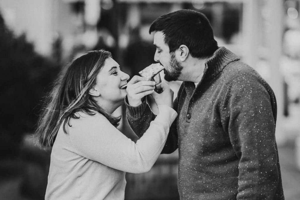 During their winter Mystic engagement session, a man and woman interlock arms while eating pizza in an alleyway.