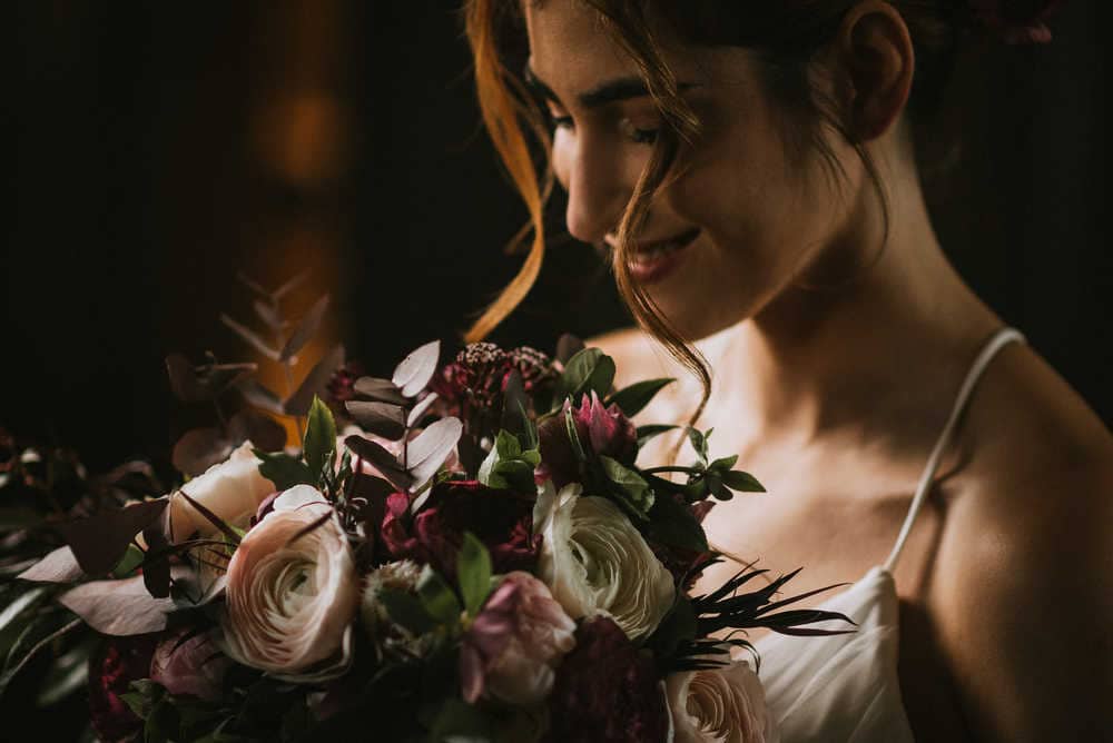 A female model at the Blithewold Mansion poses with a bridal bouquet featuring pink, white, and red flowers with green accents. Two strands of brunette hair fall on either side of her face.