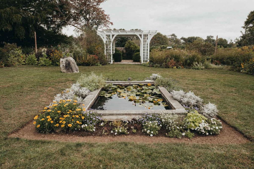 A white pergola sits in the background and a rectangular concrete pond in the foreground of the Stone Acres Farm wedding ceremony area.