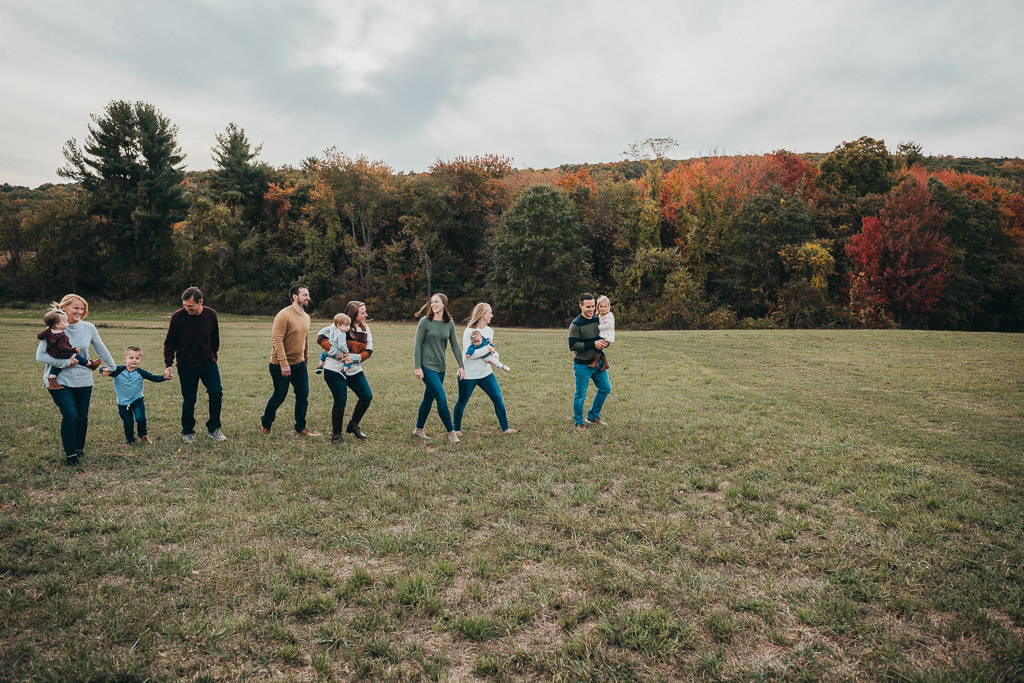 A Connecticut family during a photography session, including adults and children, walk along a grassy path in front of fall foliage and a cloudy sky.
