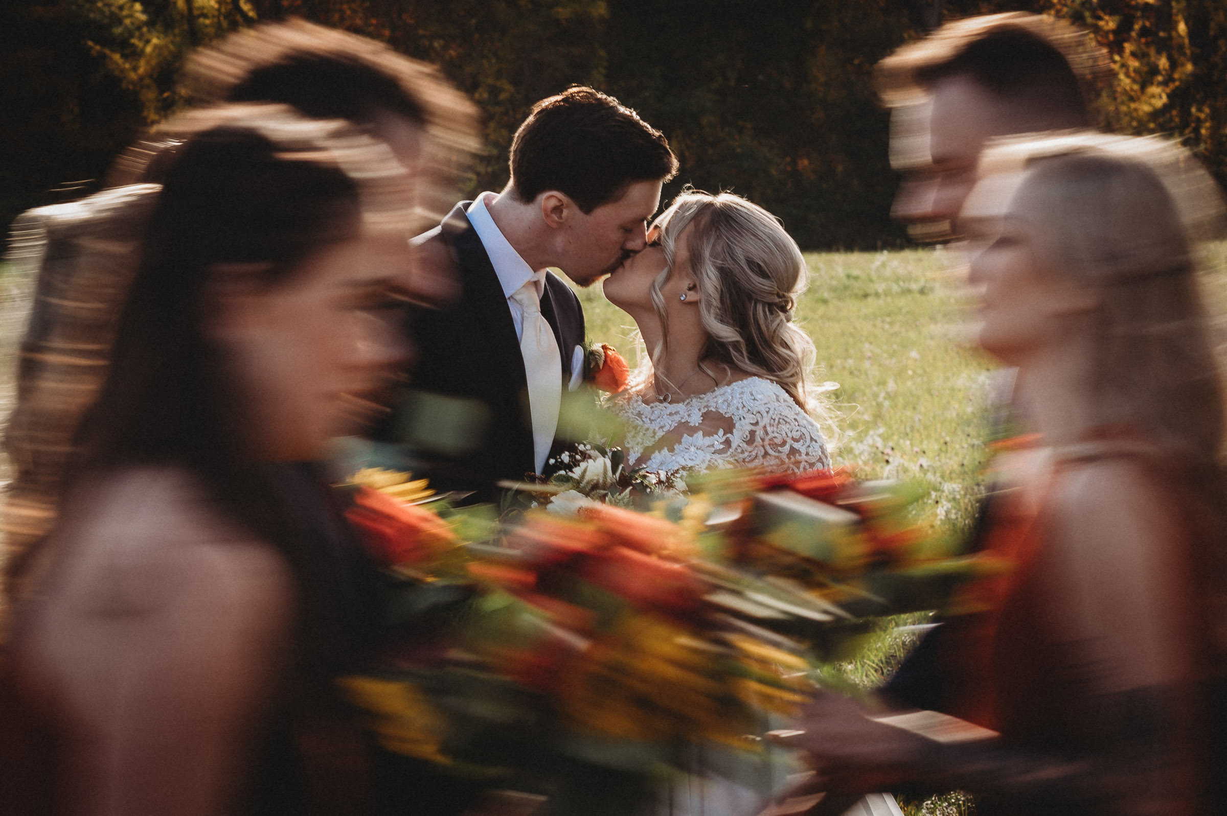 A Connecticut bride and groom kiss as their wedding party, carrying orange bouquets, walks past quickly.