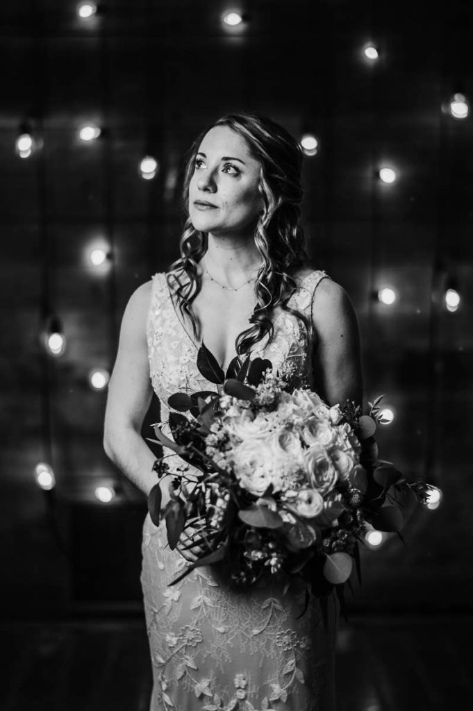 A bride poses in front of string lights on a wall before her Barns at Wesleyan Hills wedding ceremony.