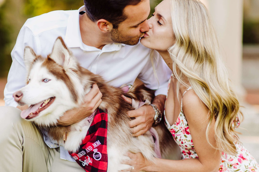 A couple enjoys a kiss while kneeling and holding their dog during their Harkness engagement session.