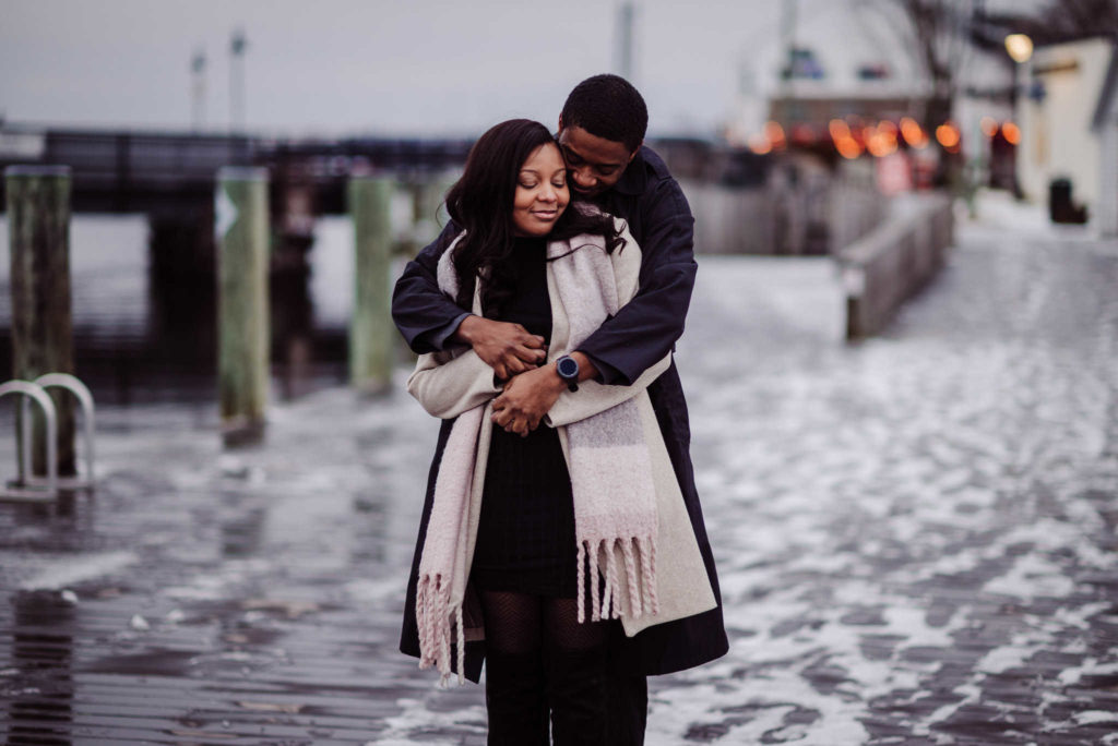 A couple stands on a dock with snow and ice for their Mystic engagement photos.