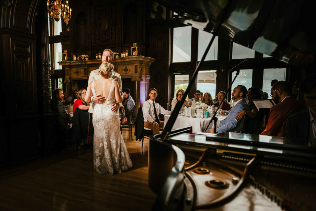 In addition to being grand and fancy, Branford House is a very affordable wedding venue in CT. Here, a bride and groom dance as a pianist sits at a grand piano and their guests watch.