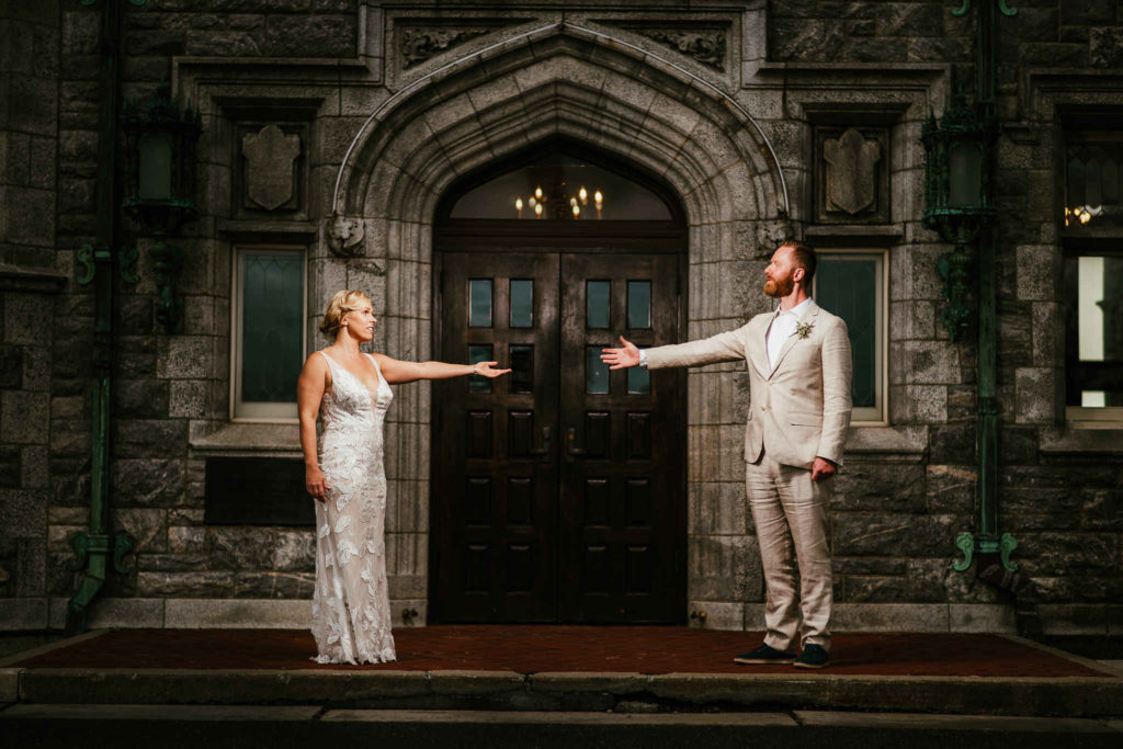 A bride and groom stand with arms outstretched in front of Branford House in Groton Connecticut.
