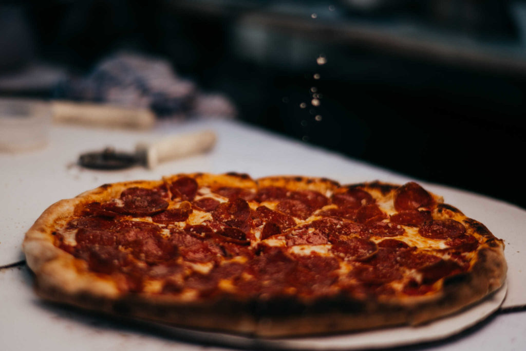 A pepperoni pizza has grated cheese sprinkled on top of it by a staff member of Victoria's, one of the best CT food trucks to rent for a wedding.