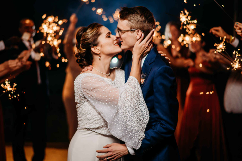 A bride and groom kiss while their wedding attendants hold orange sparklers during their Massachusetts wedding reception. Taken by Connecticut wedding photographer Terrence Irving.