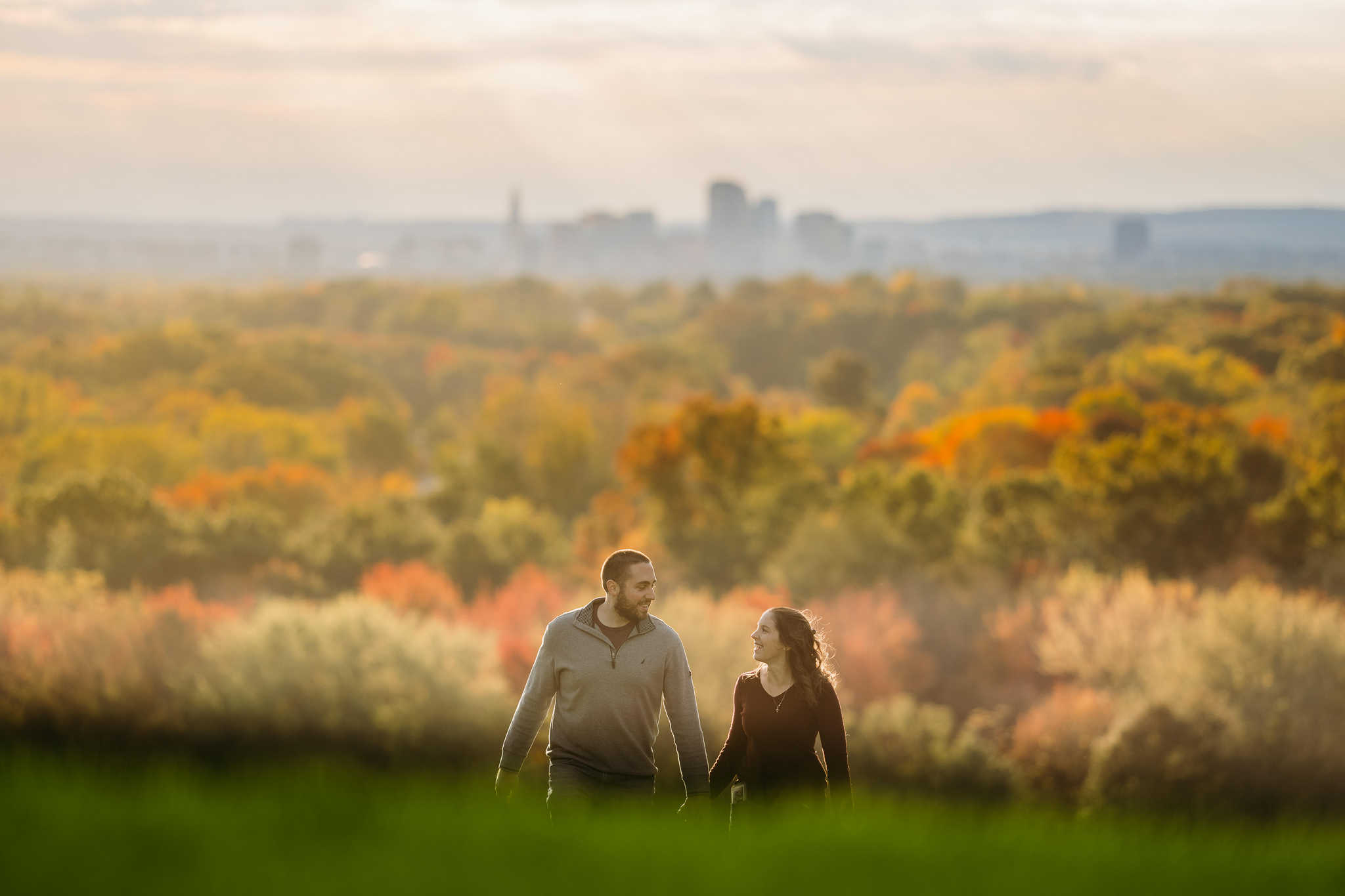 A couple holds hands and walks up a hill with the downtown Hartford skyline behind them during their Wickham Park engagement session.