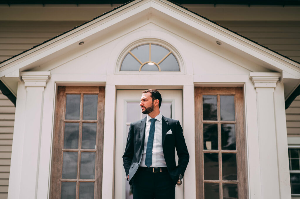 A groom stands in front of a guest house before his Lion Rock Farm wedding.