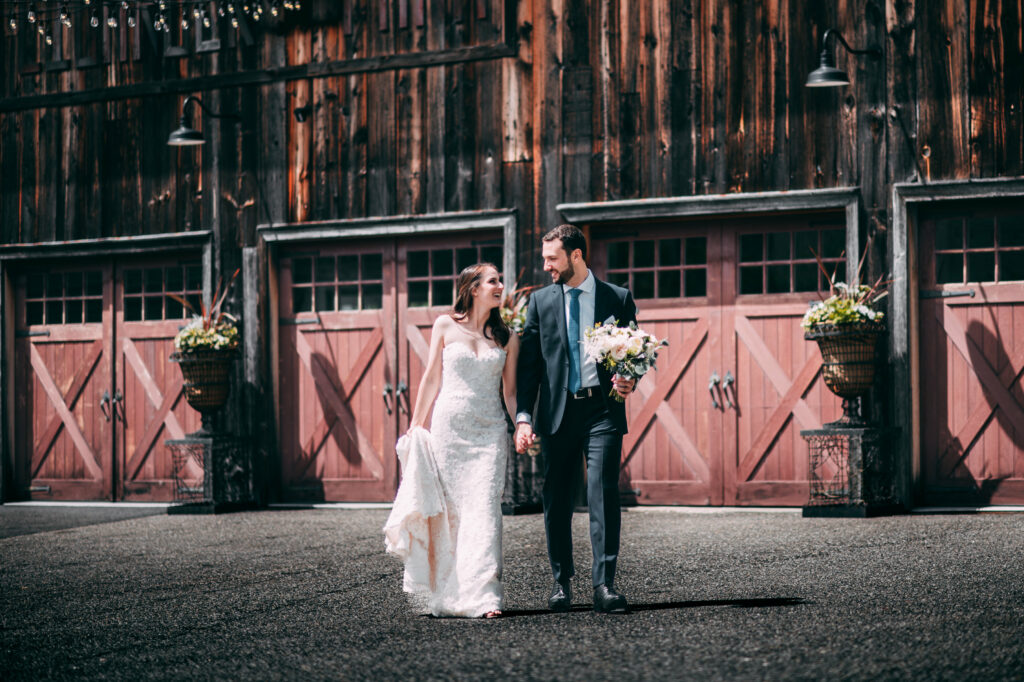 A bride and groom walk hand in hand in front of a large barn at Lion Rock Farm, an upscale rustic wedding venue in CT.