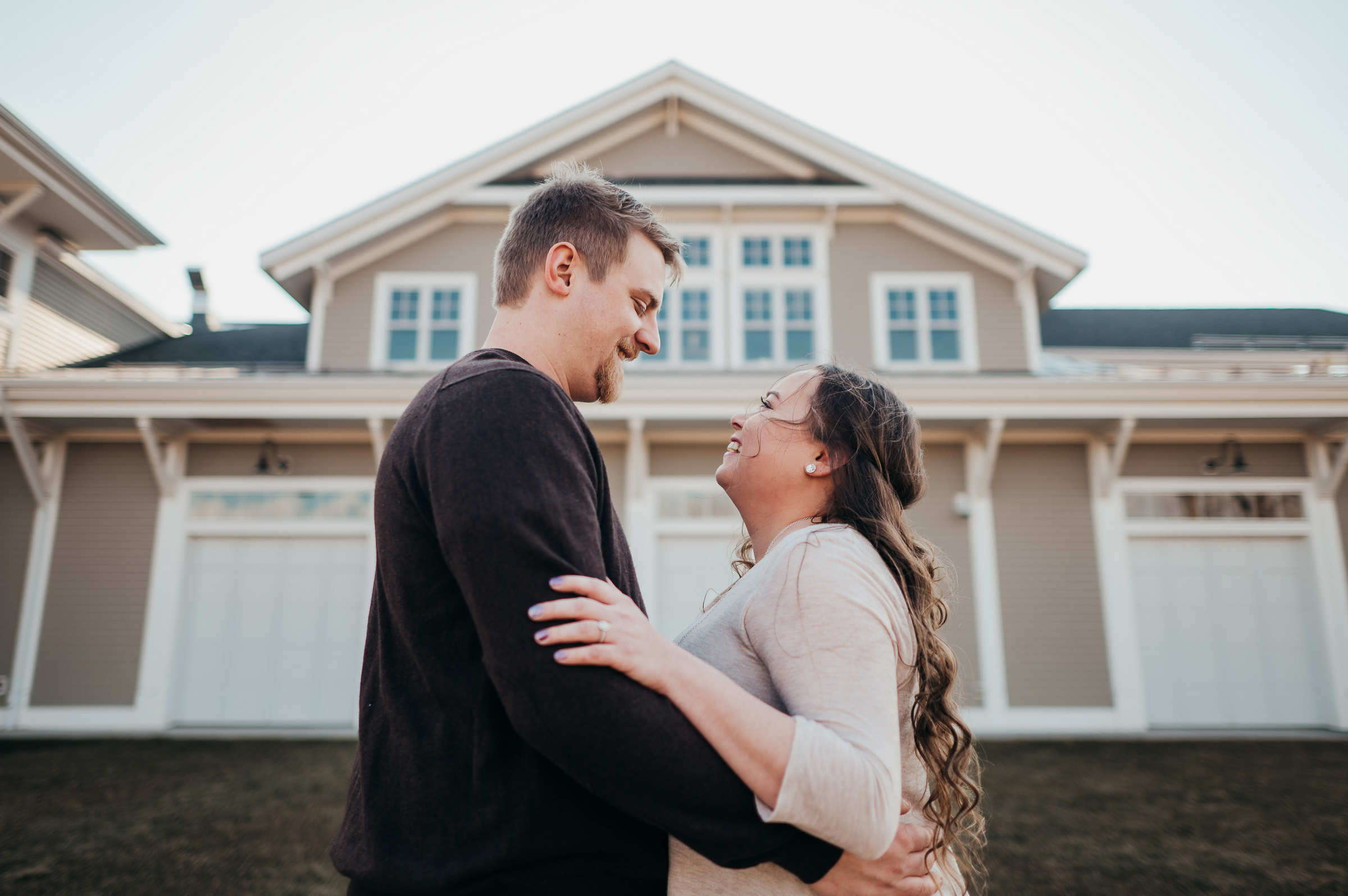 An engaged couple stands and faces each other, gently embracing and smiling, while standing in front of a boathouse during their Glastonbury engagement session.
