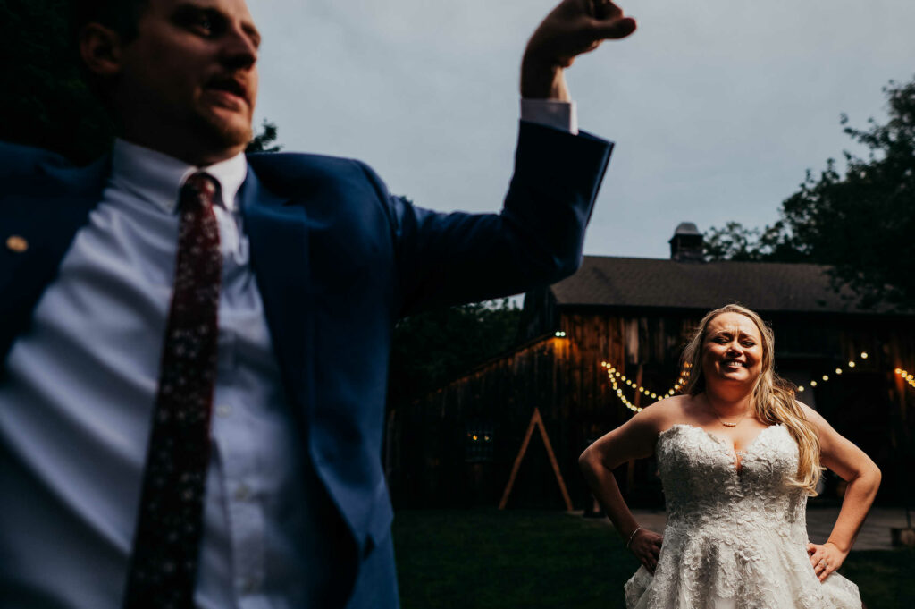 A groom flexes his muscles while his bride laughs in the background during their Webb Barn wedding.