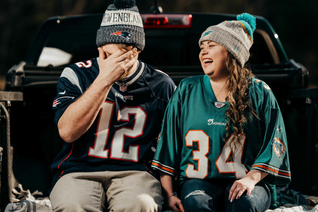 A couple wears football jerseys and hats and laughs together in a truck during their Glastonbury engagement session.