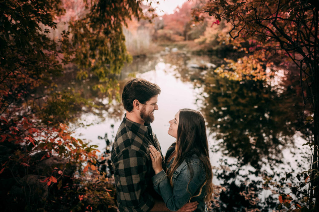 A couple stands under fall foliage in front of a pond at Haley Farm, one of the best engagement photo locations in Connecticut.