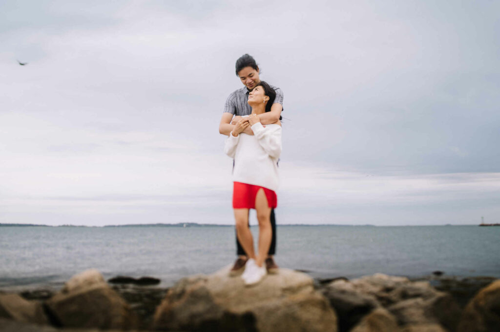 A couple embraces and stands on rocks at Stonington Point during their engagement session.