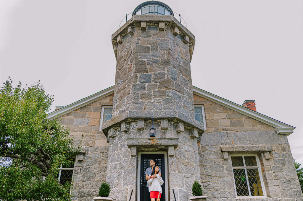 A couple poses in front of the Stonington Lighthouse Museum during their engagement session at Stonington Point.