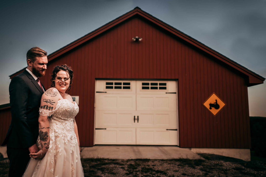 The bride and groom pose together in front of a small, red barn during their Brooklyn barn wedding.