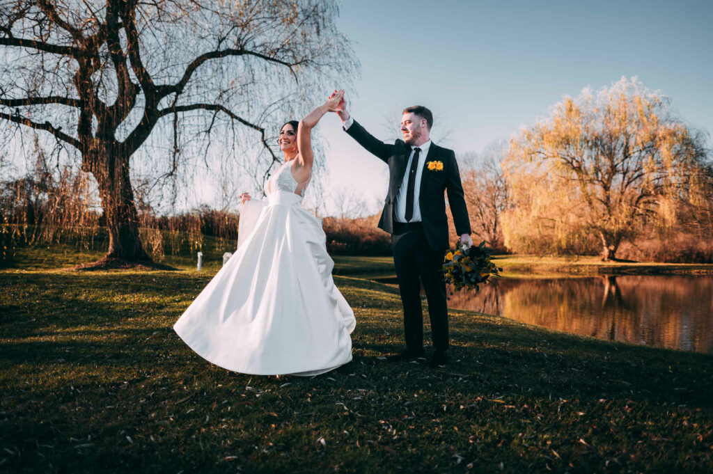 A groom twirls his bride during their wedding at The Barns at Wesleyan Hills. Willow trees and a pond are in the distance behind them.