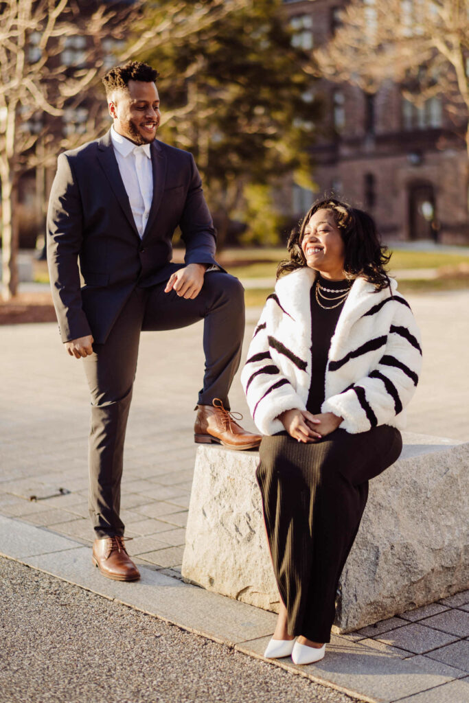 A woman sits and smiles in a fur coat while her partner smiles with his leg up on a stone block during their Trinity College engagement session in Hartford CT.