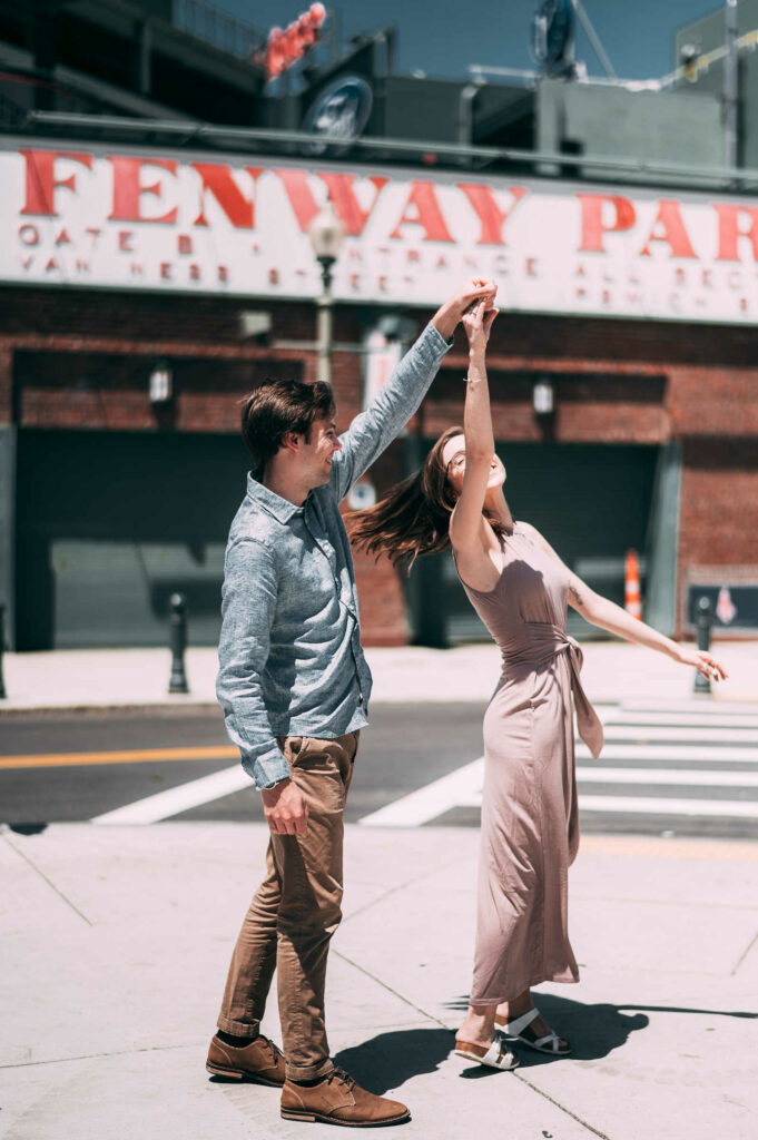 A groom-to-be twirls his fiancé in front of the Fenway stadium during their Boston engagement session.