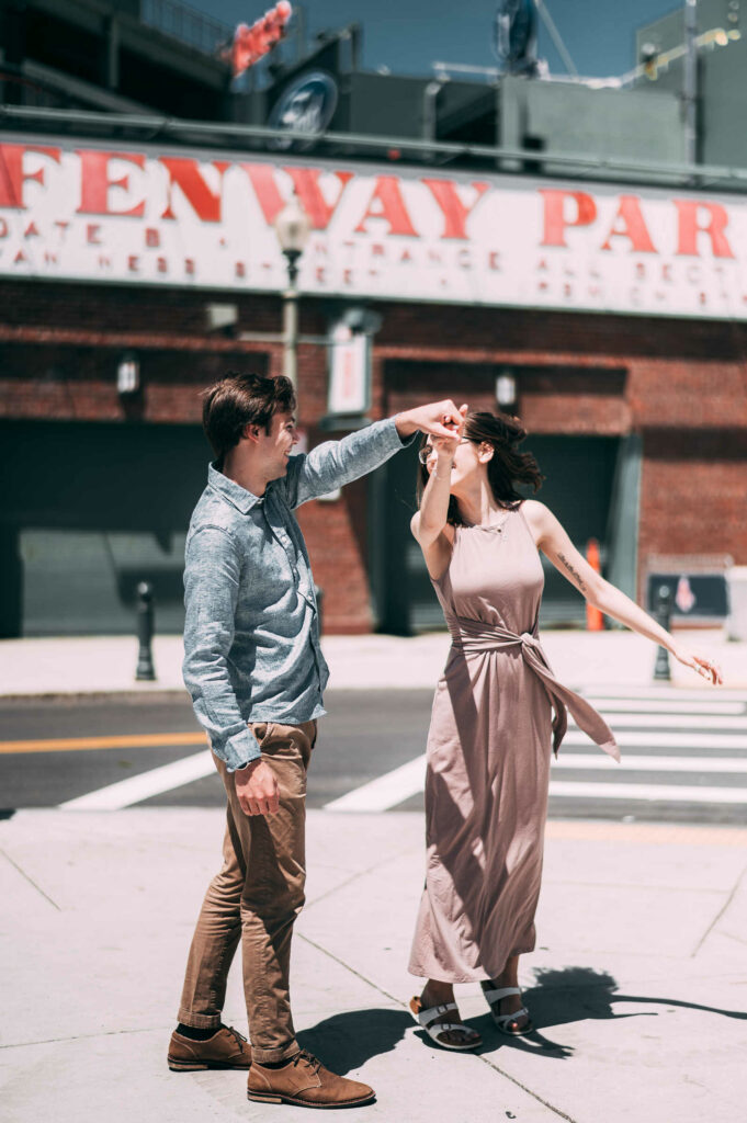 A groom-to-be twirls his fiancé in front of Fenway Park during their Boston engagement session.