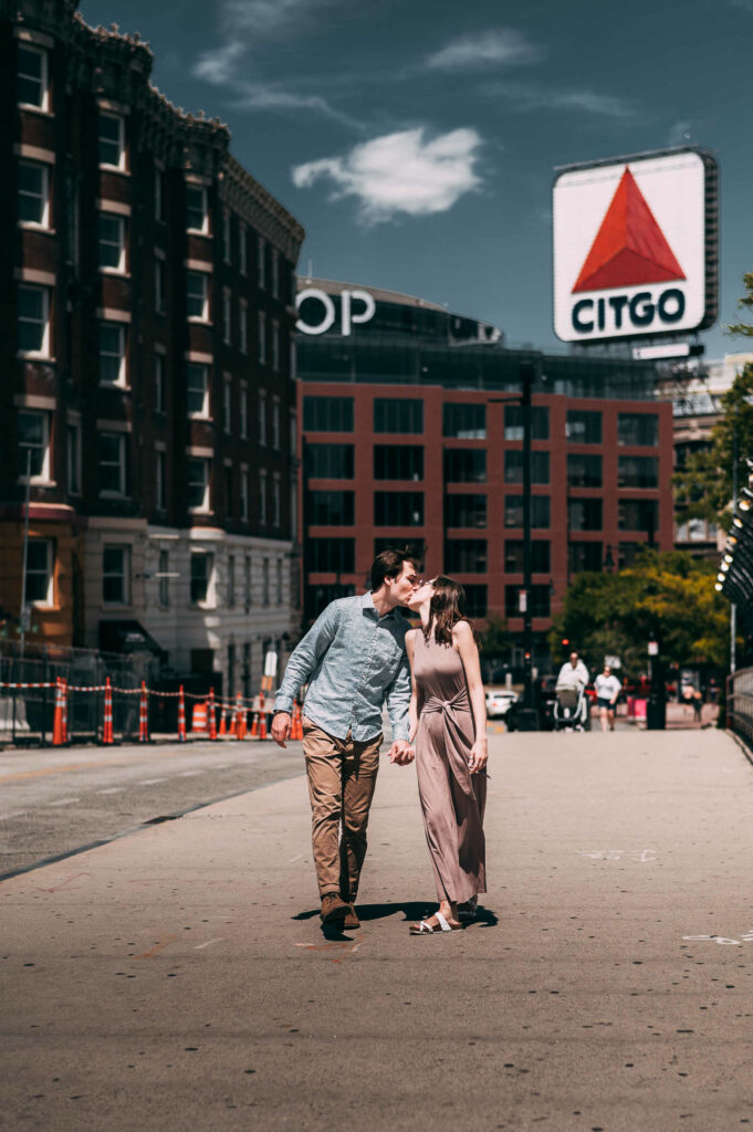 During their engagement session in Boston, an engaged couple walks together and holds hands while the Kenmore Citgo sign is in the background.