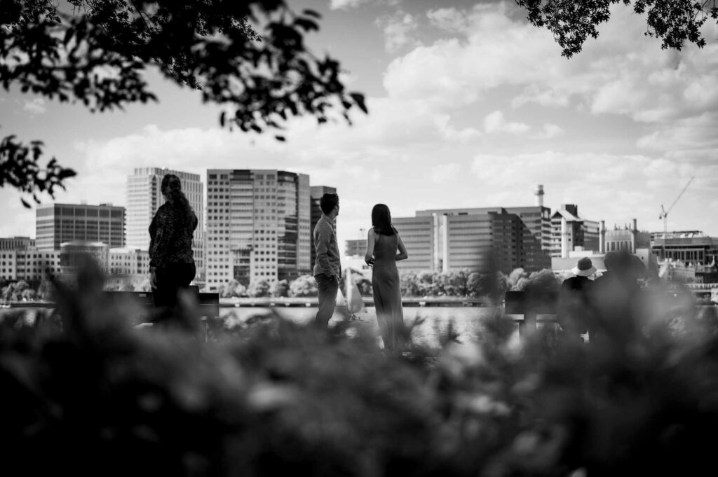 An engagement couple looks at the city skyline while standing in an esplanade during their Boston engagement session.