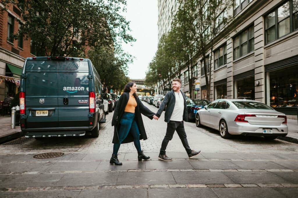 A couple holds hands and walks together towards the right, acrcoss the street past an Amazon van, during their engagement session in Brooklyn.