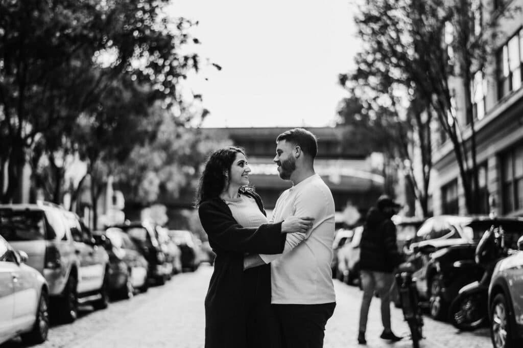 A couple embraces while standing in the middle of the street, with rows of cars parked on either side, during their engagement session in Brooklyn.