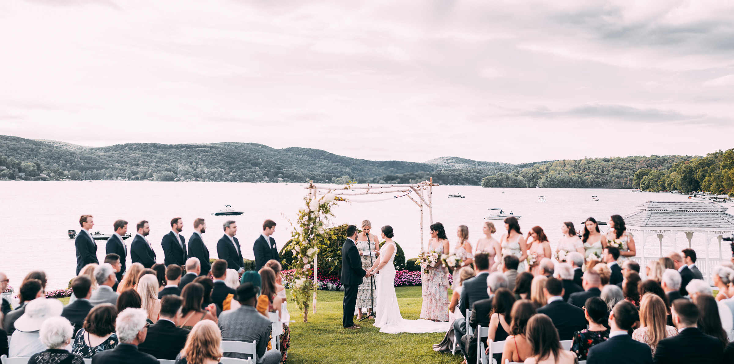 A couple stands at their altar in front of a lake, surrounded by bridesmaids in floral dresses and groomsmen in blue suits during their Candlewood Inn wedding.