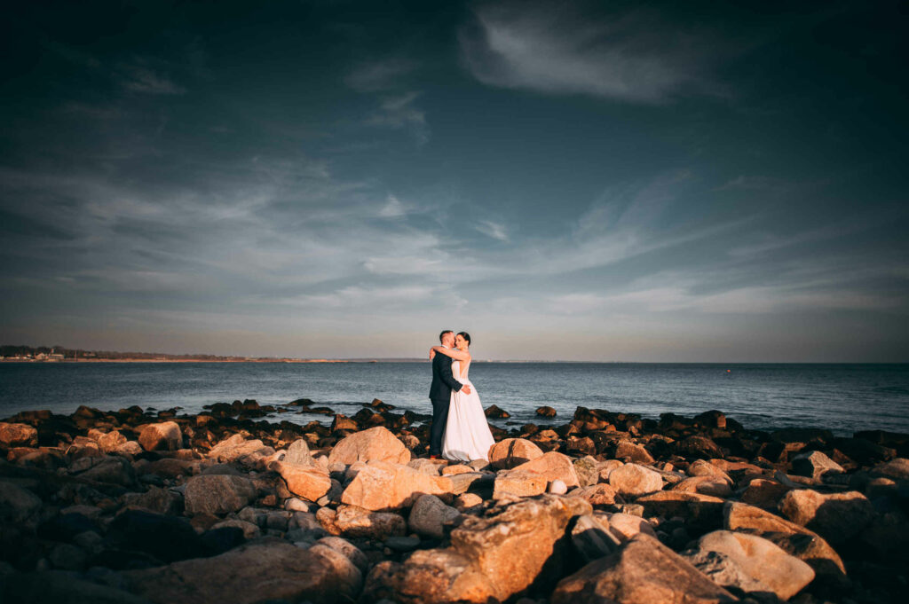 A bride and groom embrace in front of the water surrounded by rocks during their Towers Narragansett wedding.