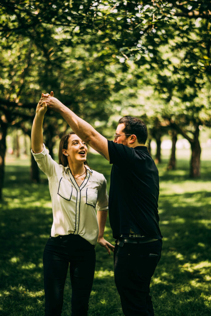 A couple dances and twirls on a green lawn surrounded by trees during their Brooklyn Botanic Garden engagement session.