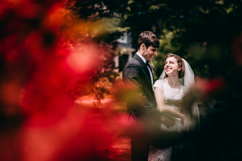 A wedding couple with classic style embrace and smile with red flowers nearby on their New Hampshire wedding day.
