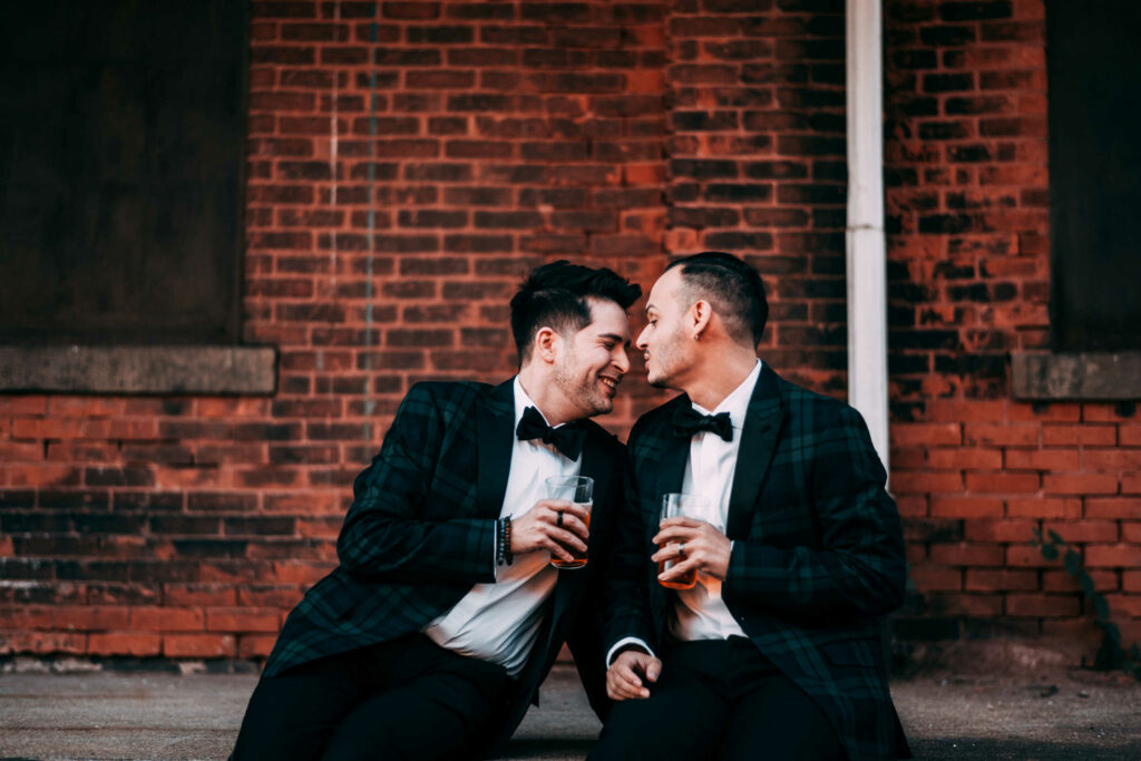 Two grooms sit next to each other in front of a brick wall while holding beers during their BADSONS brewery wedding day.