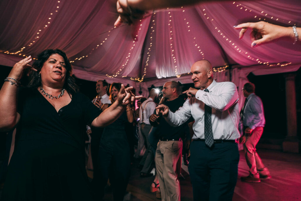 Wedding guests dance under a white tent with yellow string lights and pink uplighting during a Harkness Park wedding.
