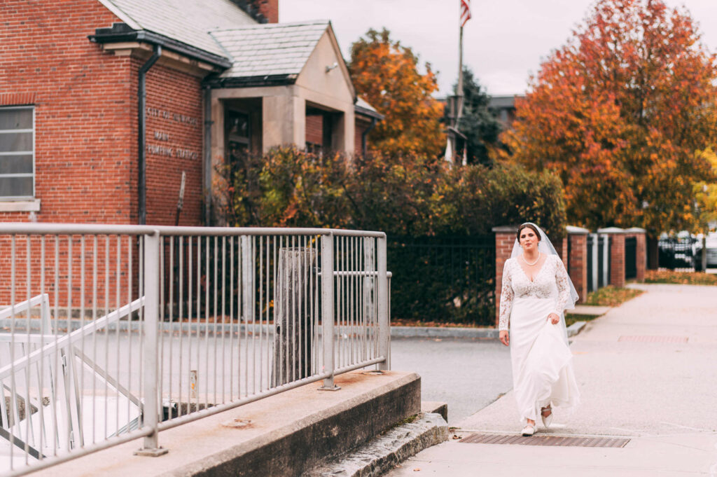 A bride in a white dress holds her train while walking down the street to the first look of her Castle Hill Inn wedding day.