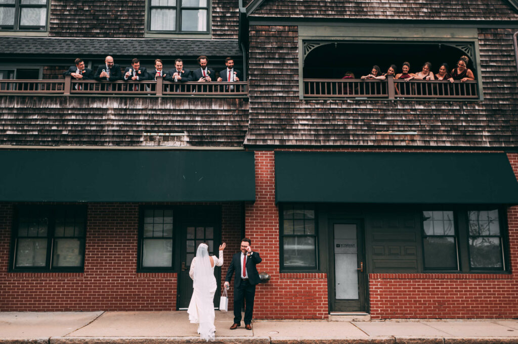 A bride and groom stand on a sidewalk by a brick wall as their wedding party members are above them on two adjoining balconies of a hotel during their Castle Hill Inn wedding day.