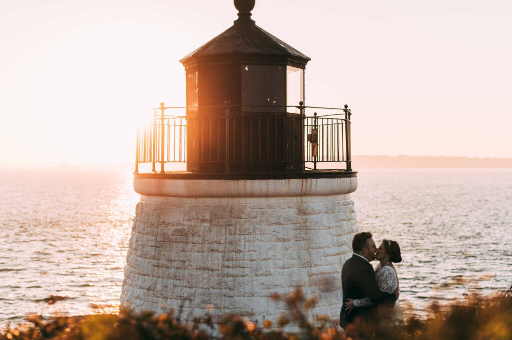 A bride and groom embrace and kiss at sunset next to Castle Hill lighthouse.
