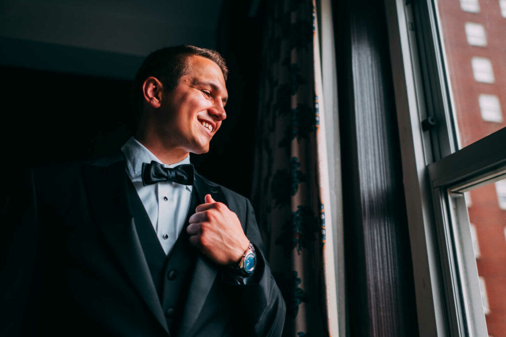 The groom poses in his black tuxedo and smiles before his wedding while in a room at Graduate Providence.