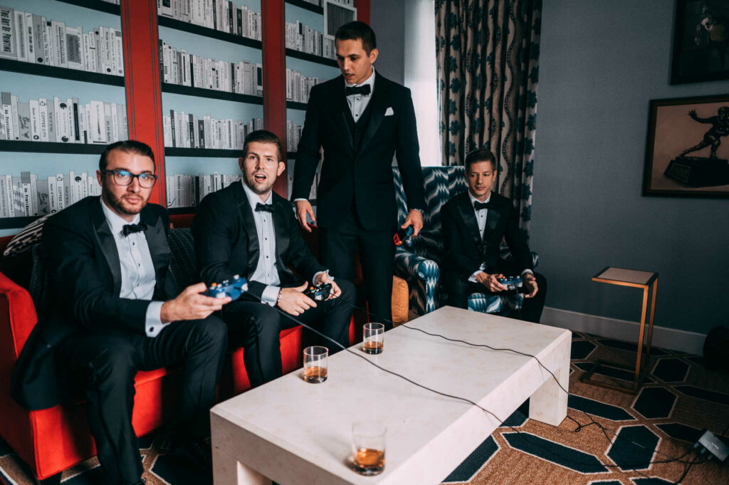Groomsmen play video games with the groom on a red couch before his Providence Biltmore wedding day.