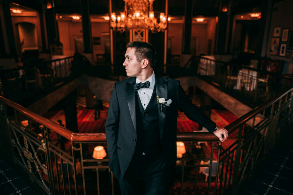 The groom has his tuxedo jacket open and leans against a railing in the mezzanine of The Biltmore on his wedding day.