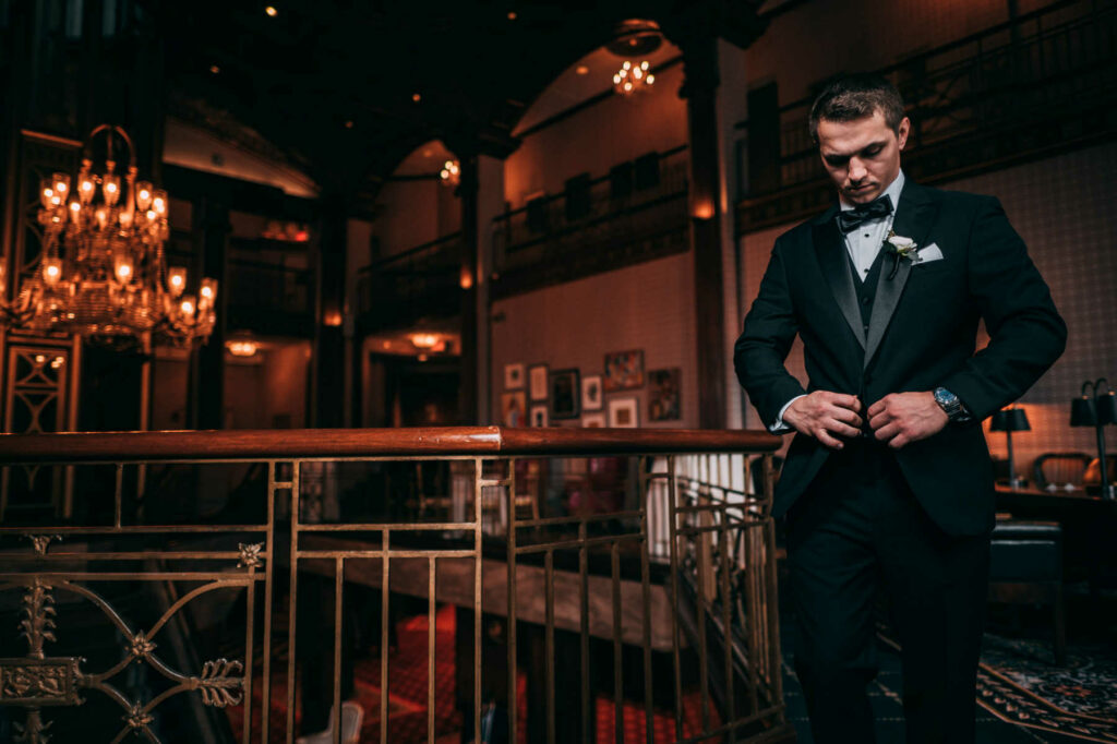 The groom in a black tux walks and buttons his jacket on the mezzanine before his wedding at Providence Biltmore.