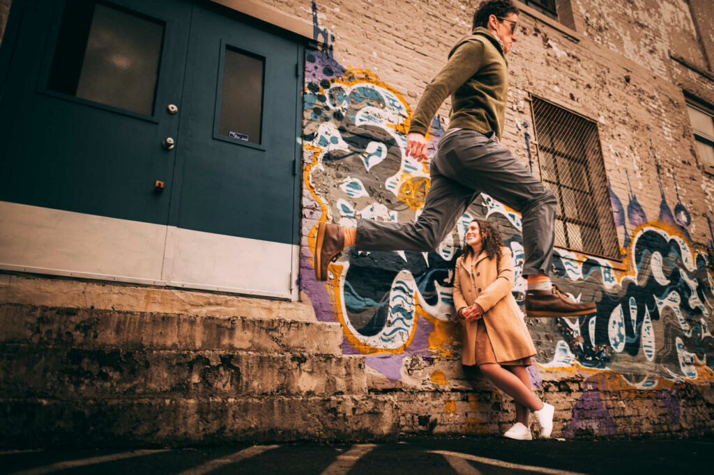 A couple jumps and smiles during their Providence engagement session in Federal Hill with a blue door and graffiti in the background.